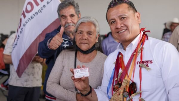 Carlos Torres Piña hablando ante público en Morelia durante evento conmemorativo del Día de las Abuelas y los Abuelos, con bandera de México y logo de programas de bienestar