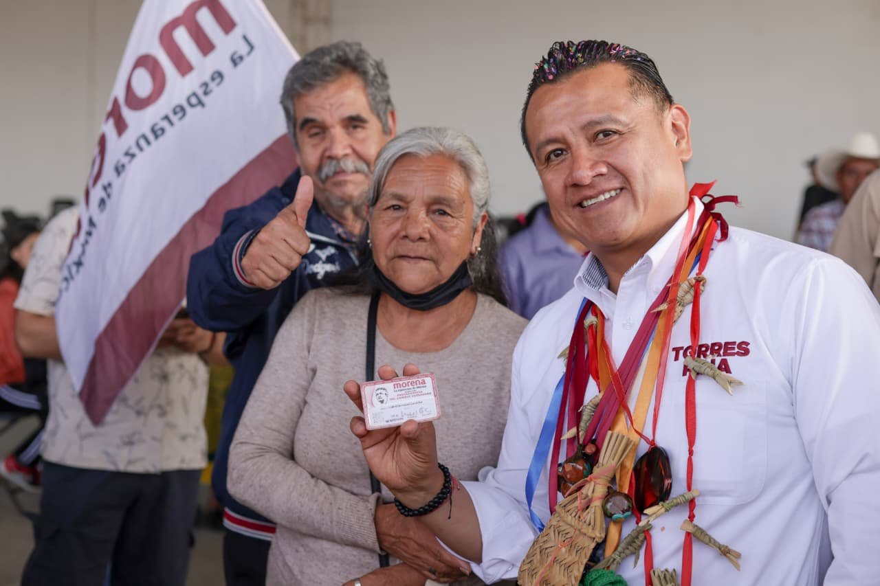 Carlos Torres Piña hablando ante público en Morelia durante evento conmemorativo del Día de las Abuelas y los Abuelos, con bandera de México y logo de programas de bienestar