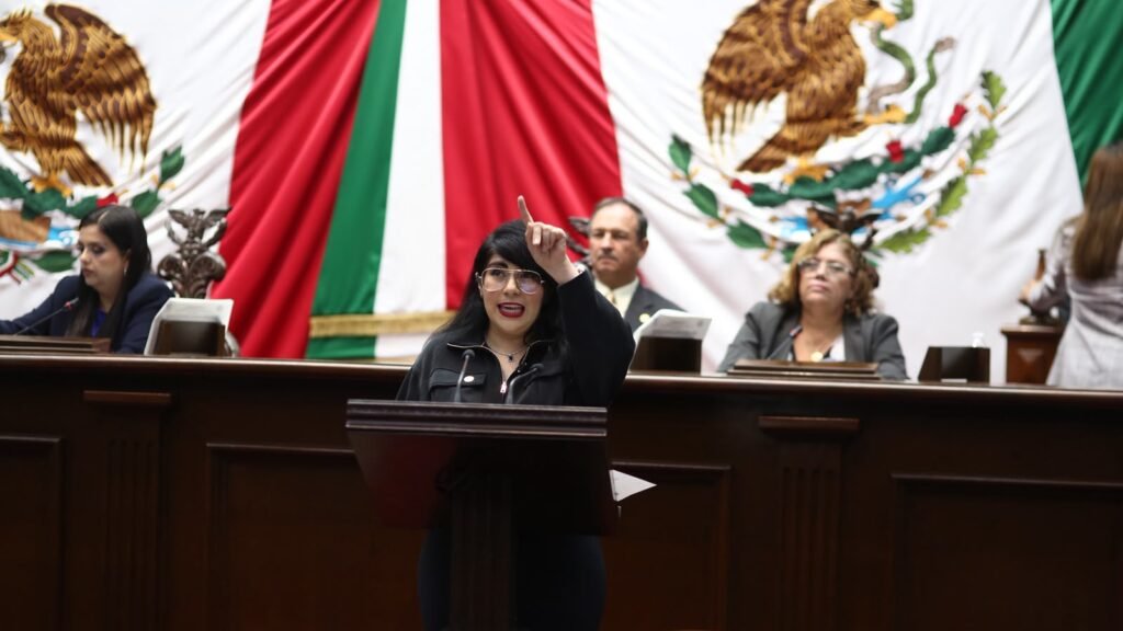 Retrato formal de la diputada Ana Vanessa Caratachea Sánchez hablando en tribuna del Congreso de Michoacán, con bandera estatal al fondo