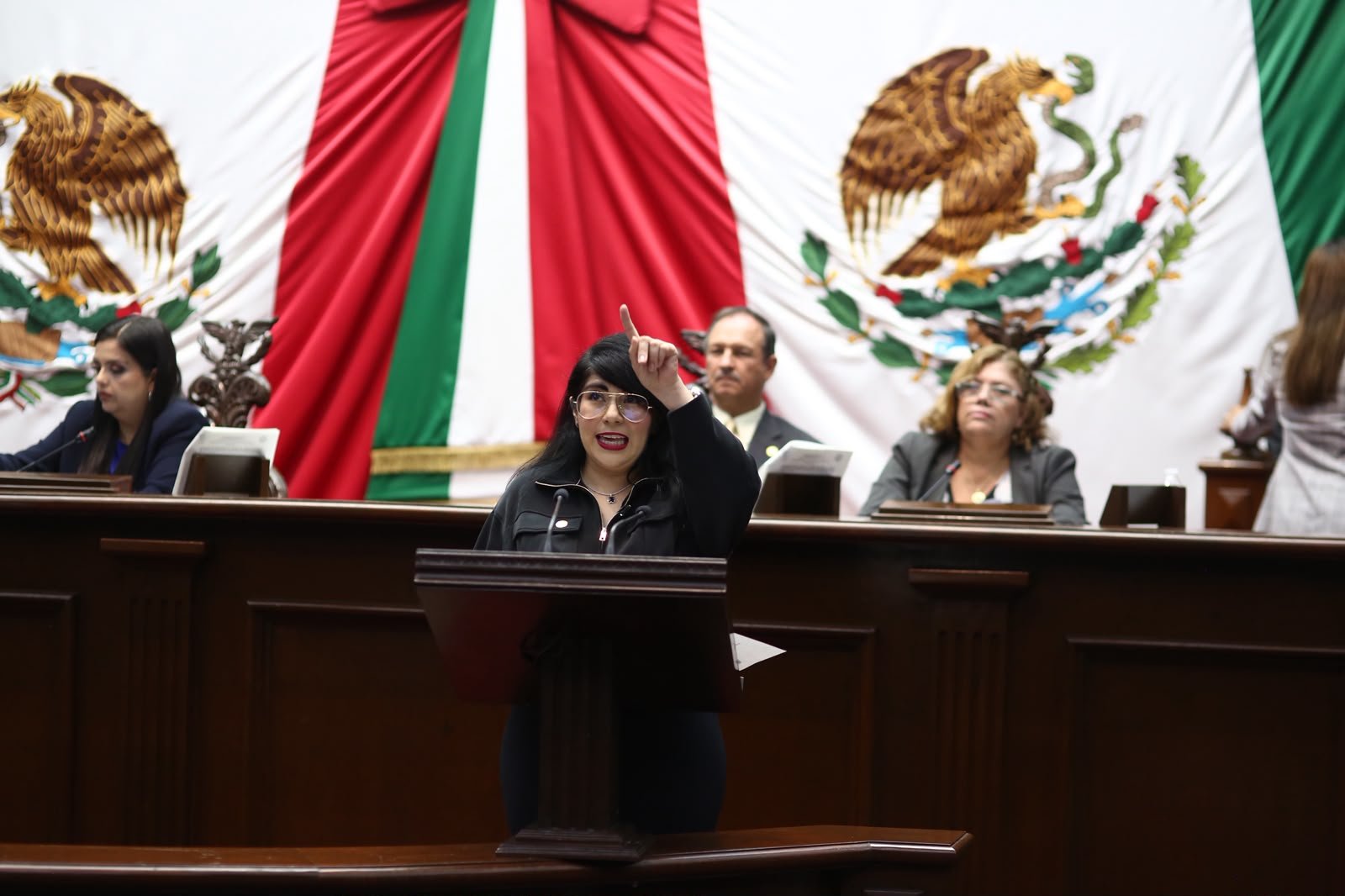 Retrato formal de la diputada Ana Vanessa Caratachea Sánchez hablando en tribuna del Congreso de Michoacán, con bandera estatal al fondo