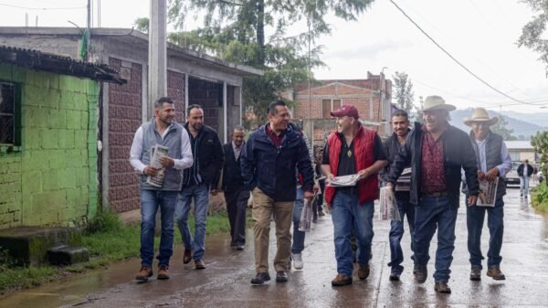 Fotografía de Carlos Torres Piña caminando por una calle rural de Michoacán, con gente local al fondo, bajo el sol, representando su recorrido constante por el territorio desde 1999