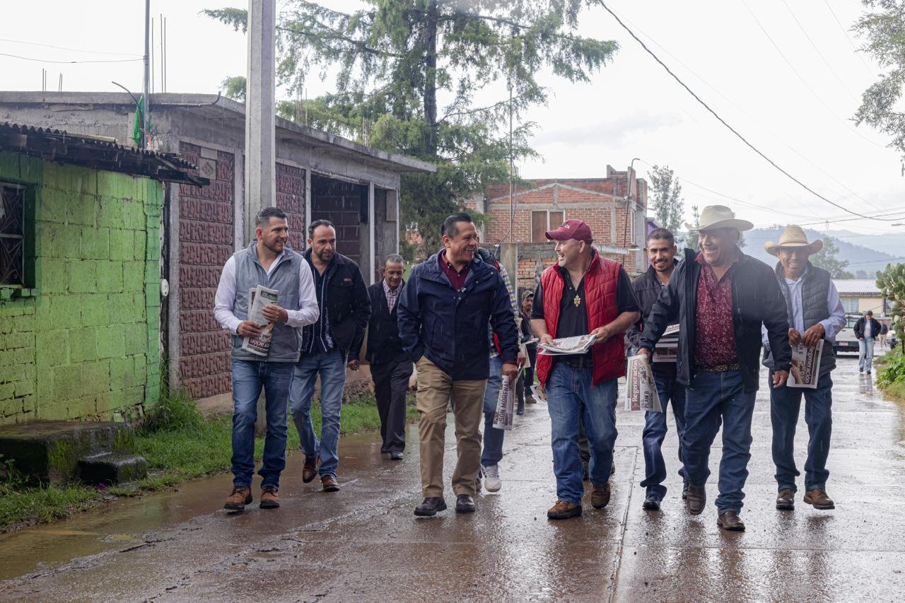 Fotografía de Carlos Torres Piña caminando por una calle rural de Michoacán, con gente local al fondo, bajo el sol, representando su recorrido constante por el territorio desde 1999
