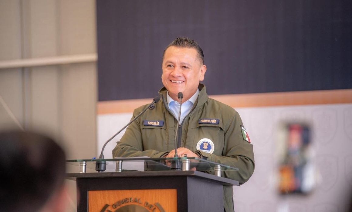 Retrato formal de Carlos Torres Piña frente a un fondo con los logos de la Fiscalía General de Michoacán y el escudo estatal
