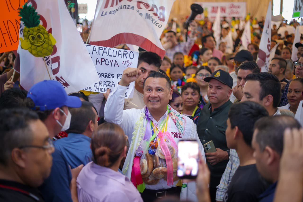 Carlos Torres Piña en una asamblea comunitaria en Michoacán, escuchando a ciudadanos en un espacio abierto, con fondo rural y presencia de personas de distintas edades.