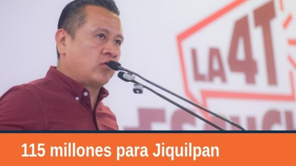 Carlos Torres Piña hablando en un evento público en Jiquilpan, Michoacán, con micrófono y fondo de bandera mexicana